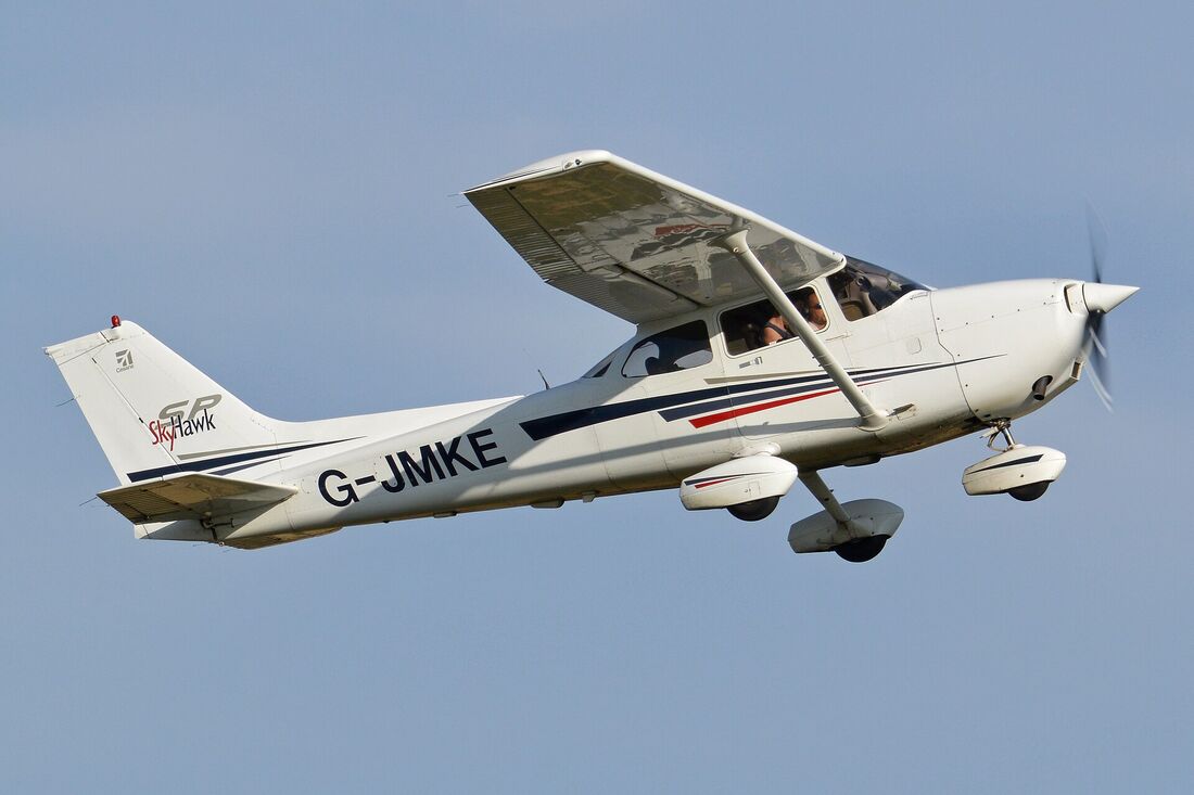 A white and blue Cessna 172 Skyhawk in flight seen from the side, with its high wing, supporting strut and fixed landing gear