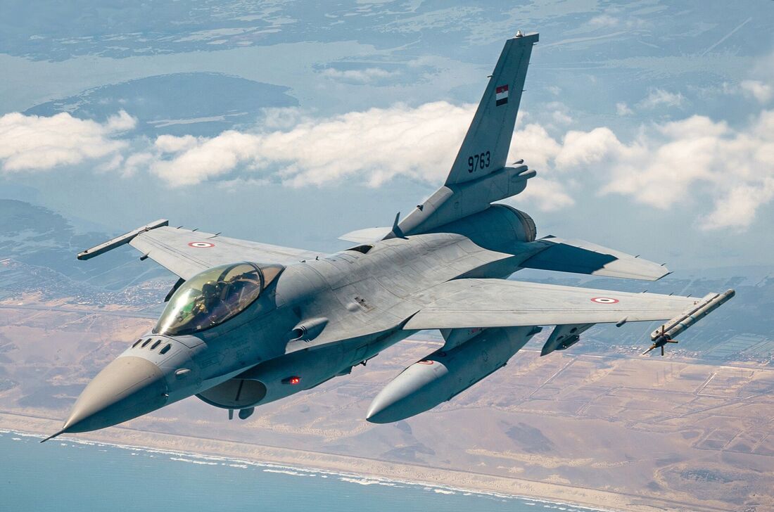 An F-16 fighter in grey camouflage banking in flight, showing the single air intake beneath its nose and a bubble canopy