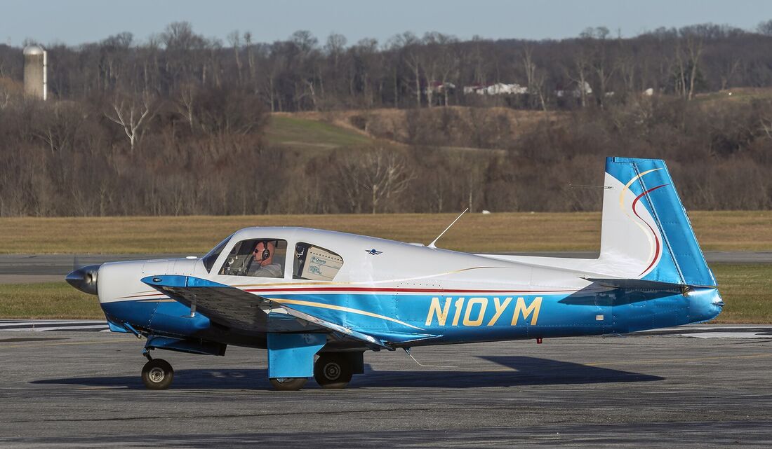 A blue and white Mooney M20 on a taxiway seen from the side, its vertical tail fin leaning distinctly forward