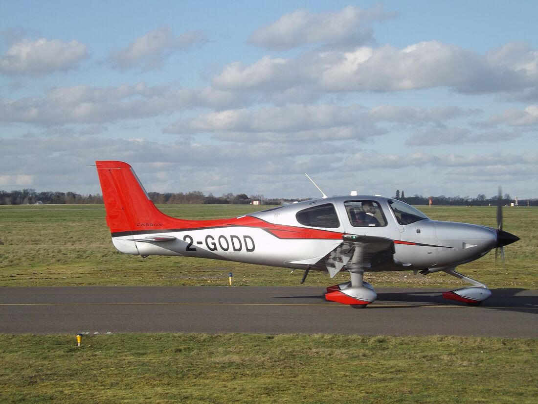A red and white Cirrus SR22 on an airfield apron, seen from the side, showing its low composite wing and swept tail fin